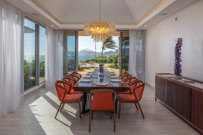 Comedor con una larga mesa dispuesta para una comida, ventanas amplias con vista al océano y una araña.