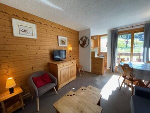 A cozy cabin interior with wooden walls, a television on a cabinet, a dining area with a table and chairs, and a large window with curtains. - Ski-in/ski-out & foot of the slopes, 1st floor, balcony, tv, ski locker, 32m², Valmorel (Valmorel)