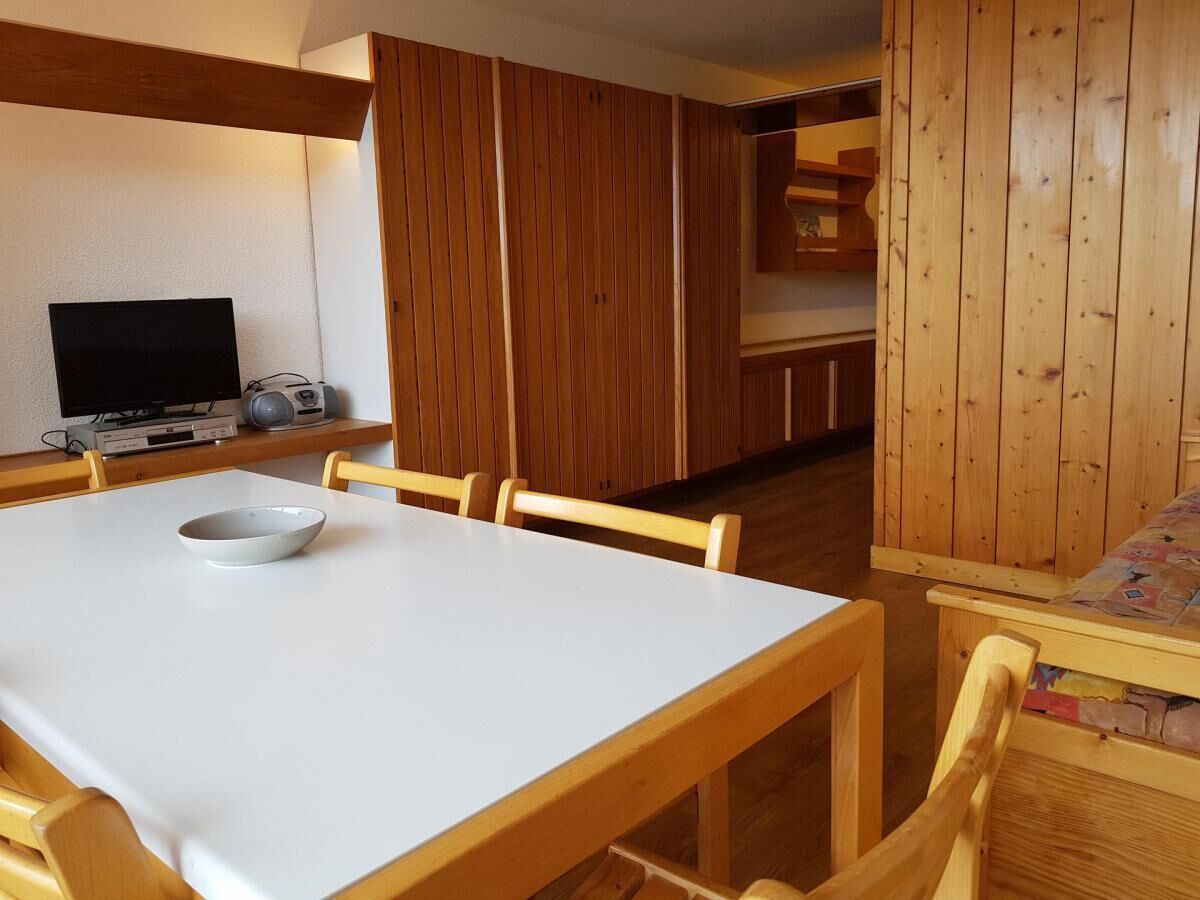 Un comedor con una mesa blanca y sillas de madera, un armario de madera con mantel acolchado, y un televisor sobre una estantería.