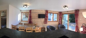 Una cabaña acogedora con interior de madera, un comedor con mesa y sillas, y un ventanal con cortinas.