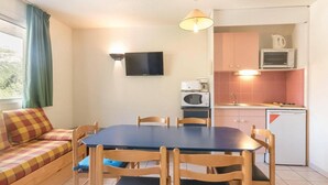 A compact living space with a dining table, chairs, a sofa, a TV, a kitchenette, and a window with curtains.