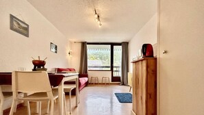A compact living space with a dining area, a sofa, a wooden cabinet, and a window with curtains. - 150m from ski slopes, 3rd floor, balcony, parking, 28m², Serre Chevalier (SERRE CHEVALIER)