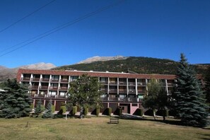 Un immeuble à plusieurs étages, doté d’un toit plat, entouré d’arbres et avec des montagnes en arrière-plan.