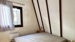 Un dormitorio con techo inclinado, una cama con colcha color crema, una ventana con cortinas y una mesita de noche pequeña.