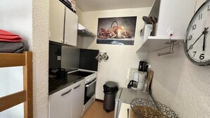 Cocina compacta con gabinetes blancos, estufa, fregadero y basurero. Hay un reloj en la pared y un estante pequeño con cafetera.