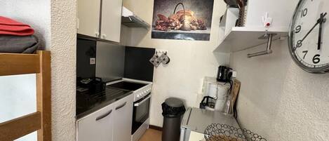 A compact kitchen with white cabinets, a stove, a sink, and a trash can. There is a clock on the wall and a small shelf with a coffee maker.