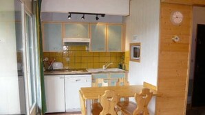 Kompakti keittiö, jossa on ruokailutila. Tilaan kuuluu puinen pöytä ja tuolit, pieni pesuallas sekä seinällä oleva kello.