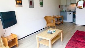 Un salon avec un téléviseur, une table en bois, un fauteuil en osier et un canapé rouge. Au fond, on aperçoit une cuisine avec des placards blancs et une horloge au mur.