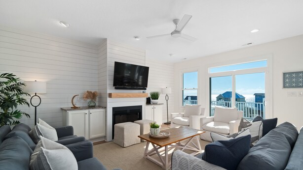 A modern living room with a fireplace, a flat-screen TV, and a large window offering a view of the ocean. - 'Coastal Vista' - Walk to Emerald Isle Beach! (Emerald Isle)