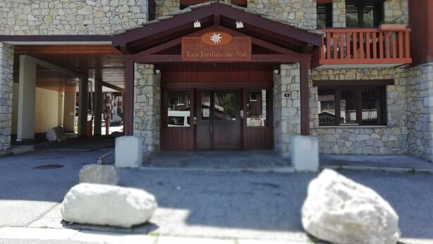 Une entrée d’immeuble avec une façade en pierre, un auvent en bois et une enseigne indiquant « Les Jardins de Vat ».