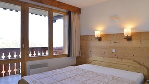 A hotel room with a wooden headboard, a bed with a white quilt, two wall-mounted lamps, and a large window with a view of the outdoors. - 100m from resort center, 5th floor, view ski slopes, balcony, tv, 39m², La Plagne (La Plagne)