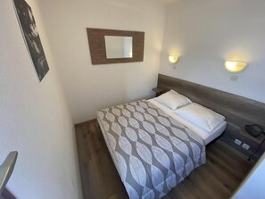 A single bed with a patterned bedspread, a wooden headboard, a wall-mounted mirror, and two wall-mounted light fixtures.