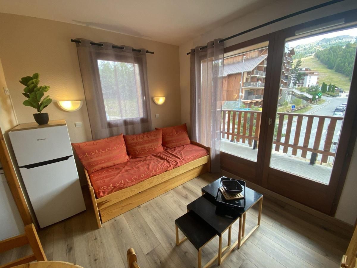 A room with a sofa bed, a small table, a refrigerator, and a view of a building through a window.