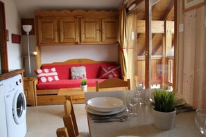 A compact room with a wooden sofa, a dining table set with plates and glasses, a washing machine, and a potted plant.