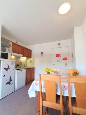 Una cocina compacta con armarios de madera, un refrigerador blanco con diseños de mariposas, un microondas y un lavavajillas. Hay una mesa de comedor con sillas y un florero con flores amarillas.