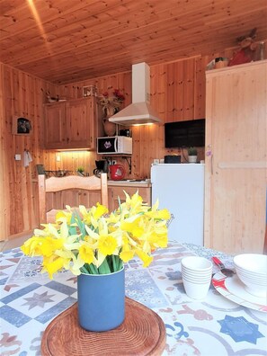 Ein Holzinterieur mit einem für zwei Personen gedeckten Esstisch, einer blauen Vase mit gelben Blumen und einer Küchenzeile mit Holzschränken und Geräten.