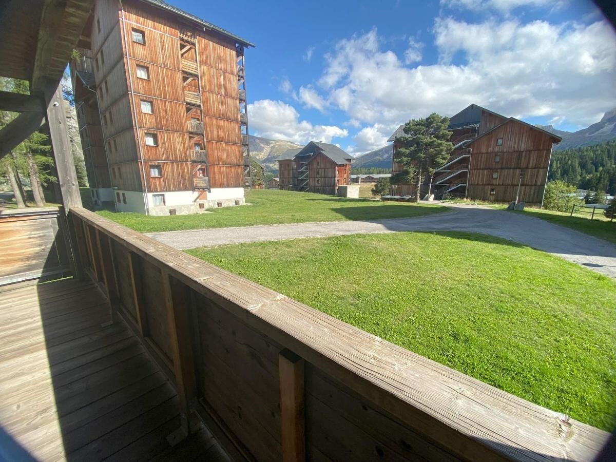 Une terrasse en bois surplombe une zone verdoyante, avec des bâtiments traditionnels en bois et des montagnes en arrière-plan.
