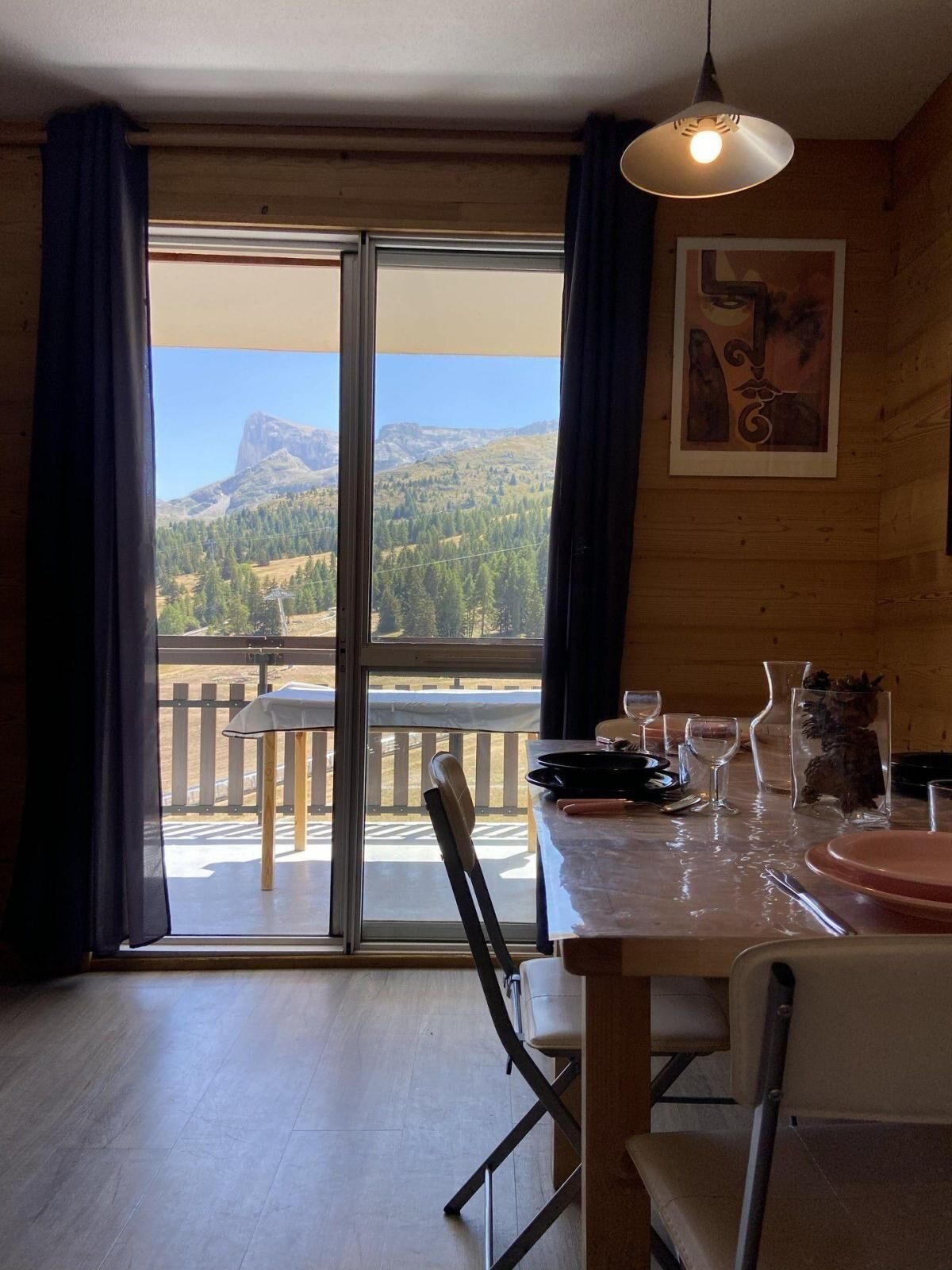 A dining area with a table set for two, a chair, a hanging light fixture, and a view of mountains through a large window.