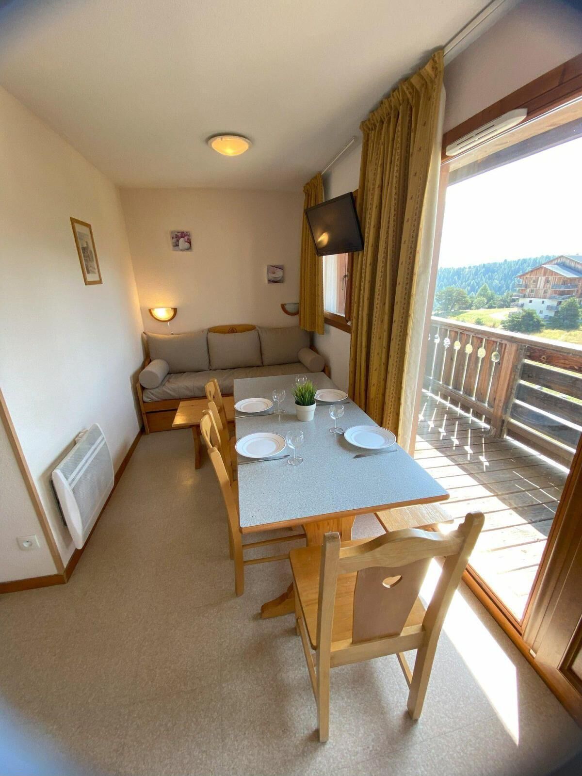 A dining area with a table set for two, a sofa, a TV, and a view of a building and trees through a window.