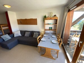 A cozy living room with a sectional sofa, a dining table with chairs, a wooden shelf with decorative items, and a sliding glass door leading to an outdoor area. - 200m from ski slopes, 2nd floor, view mountain, balcony, parking, tv, ski locker, 30m², Superdévoluy (Le Dévoluy)