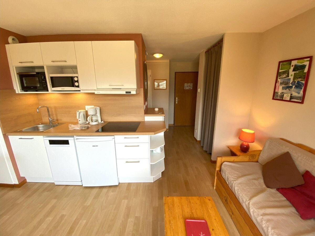 A compact hotel room with a kitchenette, a bed, a bedside table with a lamp, and a small wooden table.