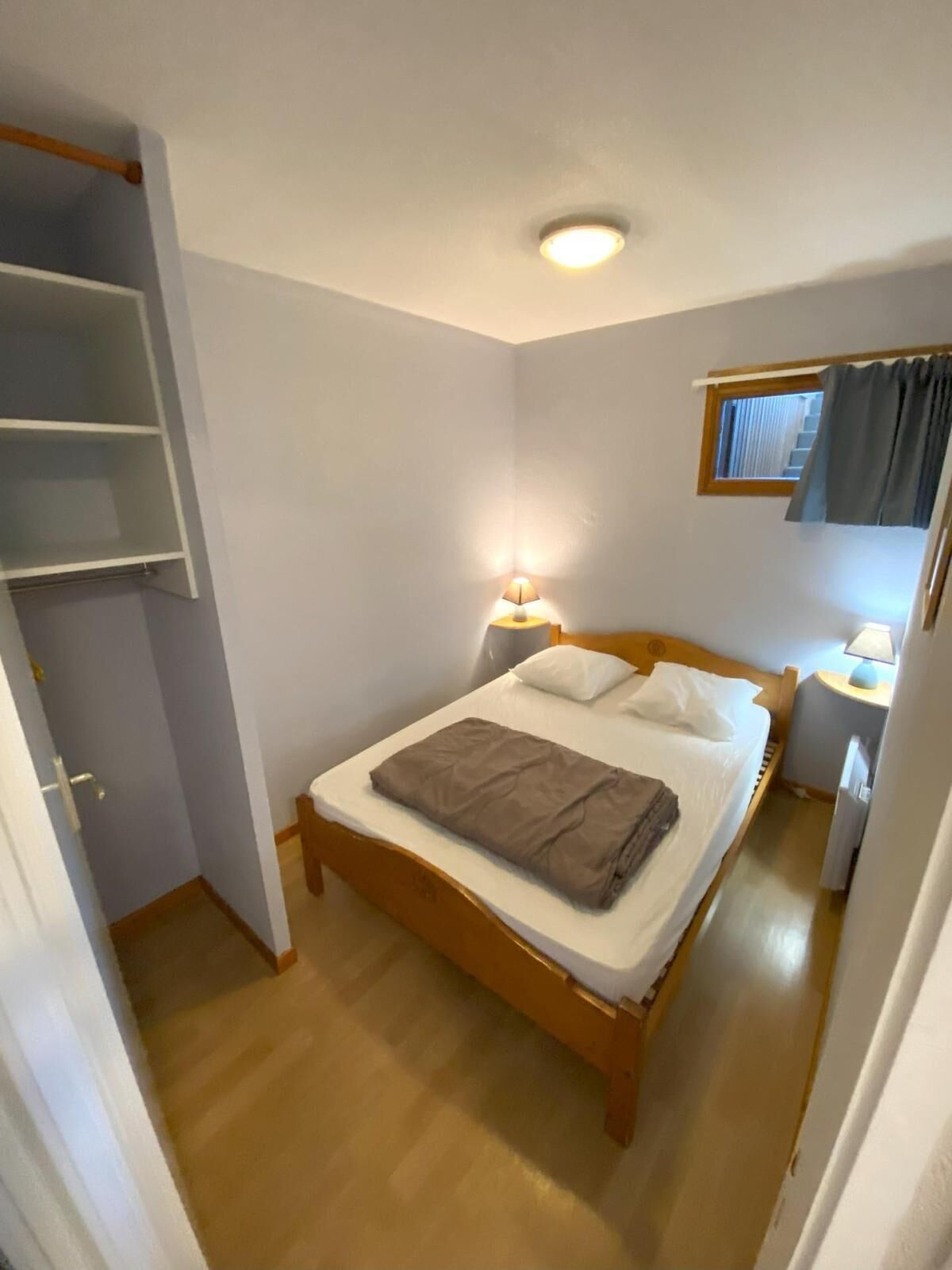 Une petite chambre à coucher, bien rangée, avec un lit simple, des tables de chevet avec des lampes, une fenêtre avec des rideaux et une armoire intégrée.