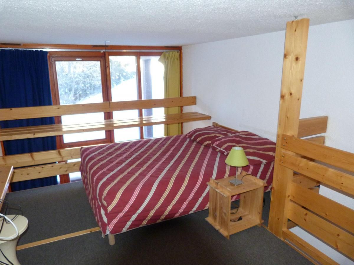 A small, single-bed room with a wooden bunk bed structure, a bedside table with a lamp, and a window with curtains.