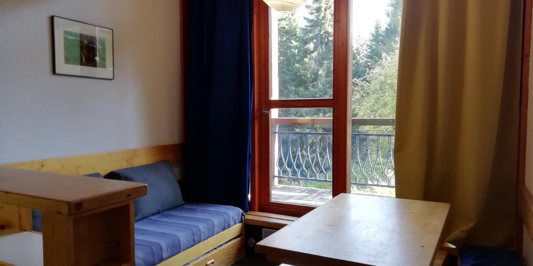 A small room with a wooden bench, a single bed, a table, and a window with a view of trees.