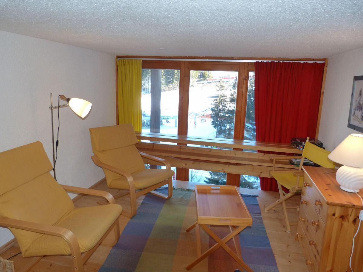 Een kamer met twee gele fauteuils, een houten tafel, een lamp en uitzicht door grote ramen met rode gordijnen.