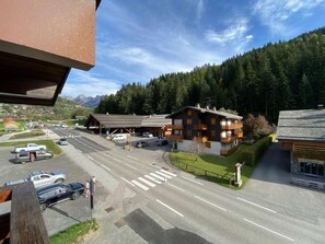 A hotel with a parking lot, a road in the foreground, and a forested mountain in the background. - 3rd floor, view mountain, balcony, tv, ski locker, 27m², Le Grand Bornand (Le Grand-Bornand)