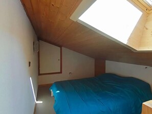 A small, single-room apartment with a sloped ceiling, a skylight, a bed with a blue cover, and a small wooden nightstand.