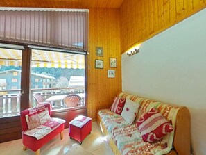 A cozy room with a wooden ceiling, a sofa, a red armchair, and a small table. There are framed pictures on the wall and a window with a view of buildings outside. - 3rd floor, view mountain, balcony, tv, ski locker, 27m², Le Grand Bornand (Le Grand-Bornand)