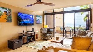 A living room with a large TV, comfortable seating, a coffee table, and a view of the ocean through large windows. -  F507 - 2 Bedroom/2 Bath - Steps from the Beach, Super Clean! (Lahaina)