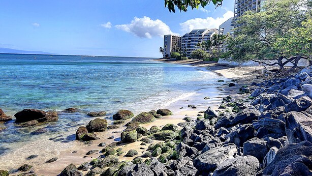 A beach with clear blue water, rocky shore, and a building with multiple floors in the background. -  F507 - 2 Bedroom/2 Bath - Steps from the Beach, Super Clean! (Lahaina)