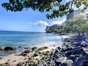 Ein Strand mit klarem, blauem Wasser, felsigem Ufer und einem mehrstöckigen Gebäude im Hintergrund.