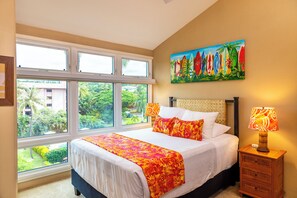 Une chambre avec une grande fenêtre, un lit recouvert d’une couverture aux couleurs vives, une table de chevet avec une lampe et un tableau accroché au mur.