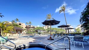 A pool area with lounge chairs and umbrellas, surrounded by palm trees and buildings. -  F507 - 2 Bedroom/2 Bath - Steps from the Beach, Super Clean! (Lahaina)