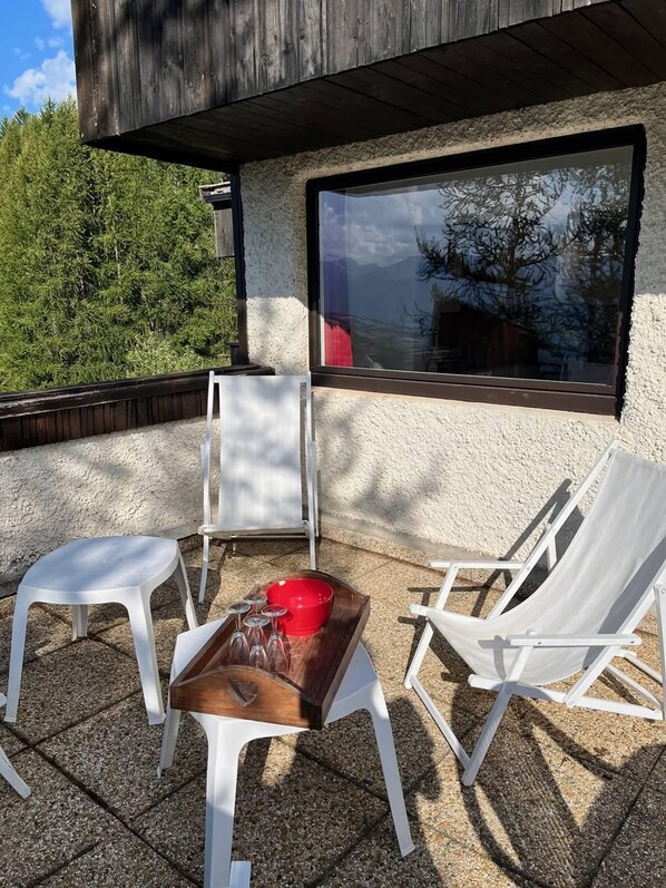 An outdoor seating area with white plastic chairs and a wooden table, a window with a view of trees, and a textured wall.