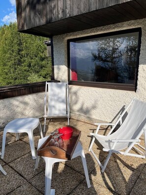 Ein Sitzbereich im Freien mit weißen Plastikstühlen und einem Holztisch, ein Fenster mit Blick auf Bäume und eine strukturierte Wand.