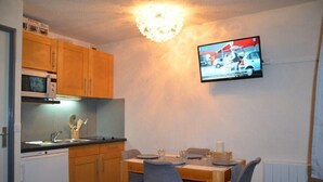Une cuisine compacte avec des meubles en bois, un four à micro-ondes, un évier et une table à manger avec des chaises. Un téléviseur à écran plat est fixé au mur, au-dessus de la table.