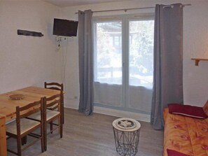Une pièce comprenant une table et des chaises en bois, une petite table ronde ornée d’un objet décoratif, un canapé avec un coussin rouge et un téléviseur fixé au mur.