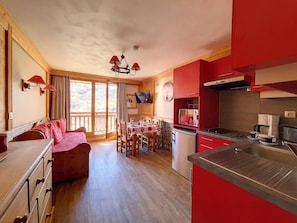 Une pièce chaleureuse avec un plancher en bois, un coin repas avec une table et des chaises, une kitchenette dotée de meubles rouges et une grande fenêtre avec des rideaux.