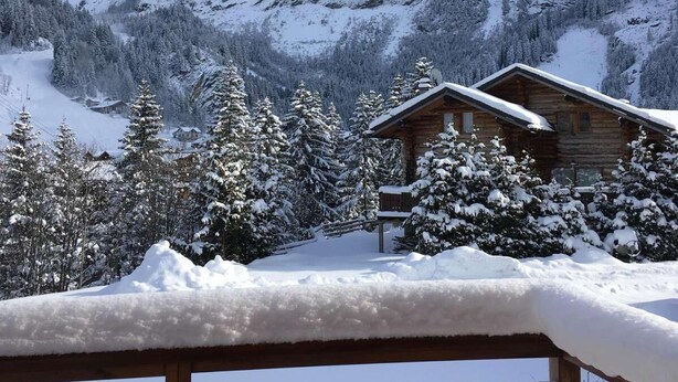 A wooden cabin with a snow-covered deck, surrounded by snow-laden trees and mountains.