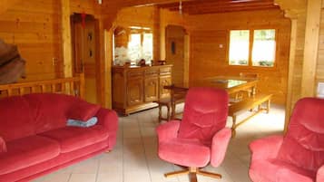 A wooden cabin interior with a red sofa, two red armchairs, a wooden dining table, and a cabinet.