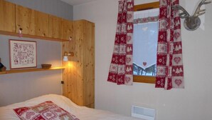 Une chambre avec une tête de lit en bois, un lit recouvert d’une literie blanche et agrémenté d’un coussin à motifs rouges, une fenêtre avec des rideaux rouges et blancs, et une tête de cerf fixée au mur.