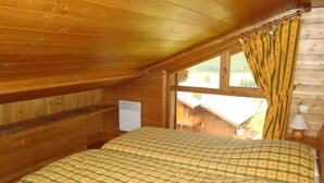 Une pièce chaleureuse avec un plafond en bois, un lit recouvert d’une courtepointe à carreaux, une fenêtre avec des rideaux et une petite table de chevet avec une lampe.