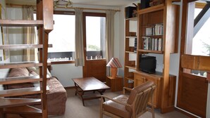 Um quarto aconchegante com um sofá de madeira, uma mesinha, uma cadeira, uma estante de livros e uma televisão.