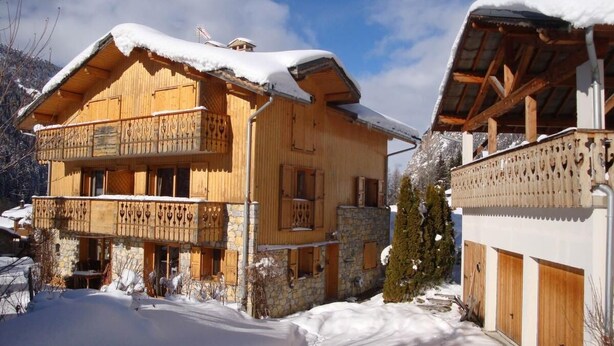 A wooden chalet with a balcony, stone foundation, and snow-covered roof.