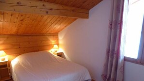 A bedroom with a wooden ceiling, a bed with white bedding, two bedside lamps, and a window with floral curtains.