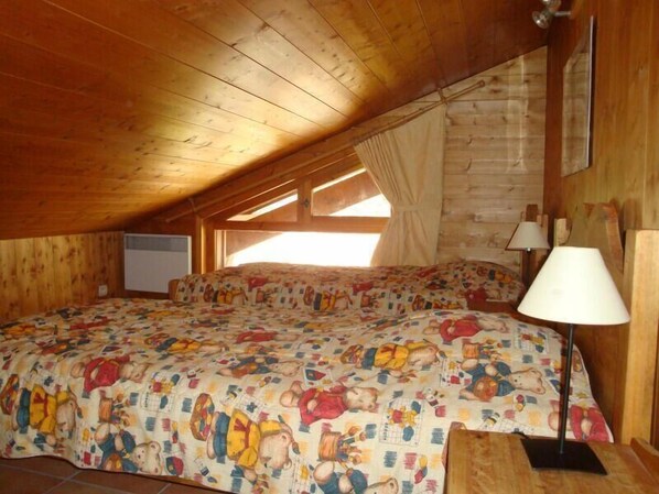 A cozy attic bedroom with wooden beams, a bed with a colorful cartoon print bedspread, a bedside lamp, and a window with curtains.